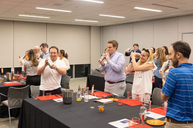 Adults in an office conference room laughing and shaking cocktail shakers during a mixology team-building workshop, tables set with bottles, tools and garnishes.