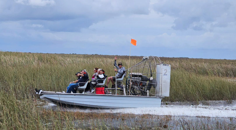 Airboat with five passengers skimming through tall marsh grasses under a cloudy sky, orange safety flag visible — Florida Everglades-style swamp tour.