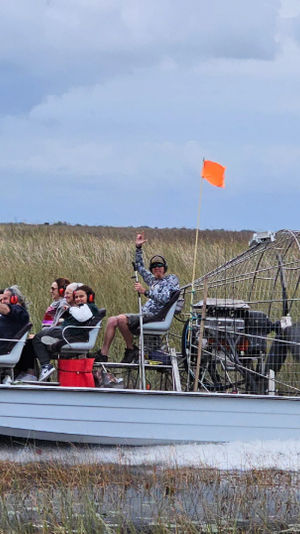Airboat with five passengers skimming through tall marsh grasses under a cloudy sky, orange safety flag visible — Florida Everglades-style swamp tour.