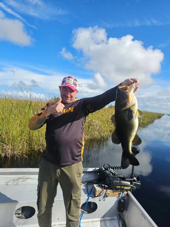 Smiling angler in an aluminum boat holding a large largemouth bass above calm freshwater marsh waters under a bright blue sky with fluffy clouds