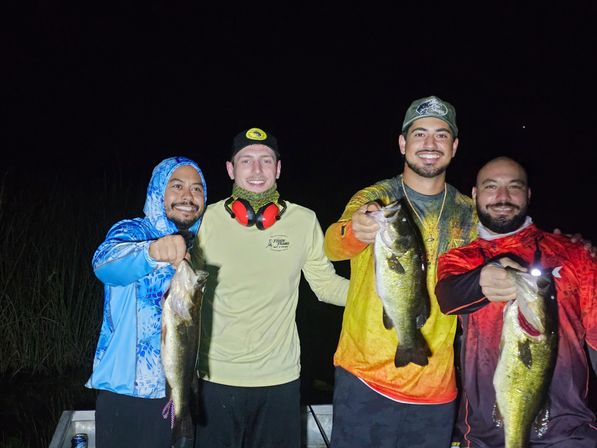 Four smiling men on a fishing boat at night holding largemouth bass, wearing colorful fishing shirts, caps and gear on a lake