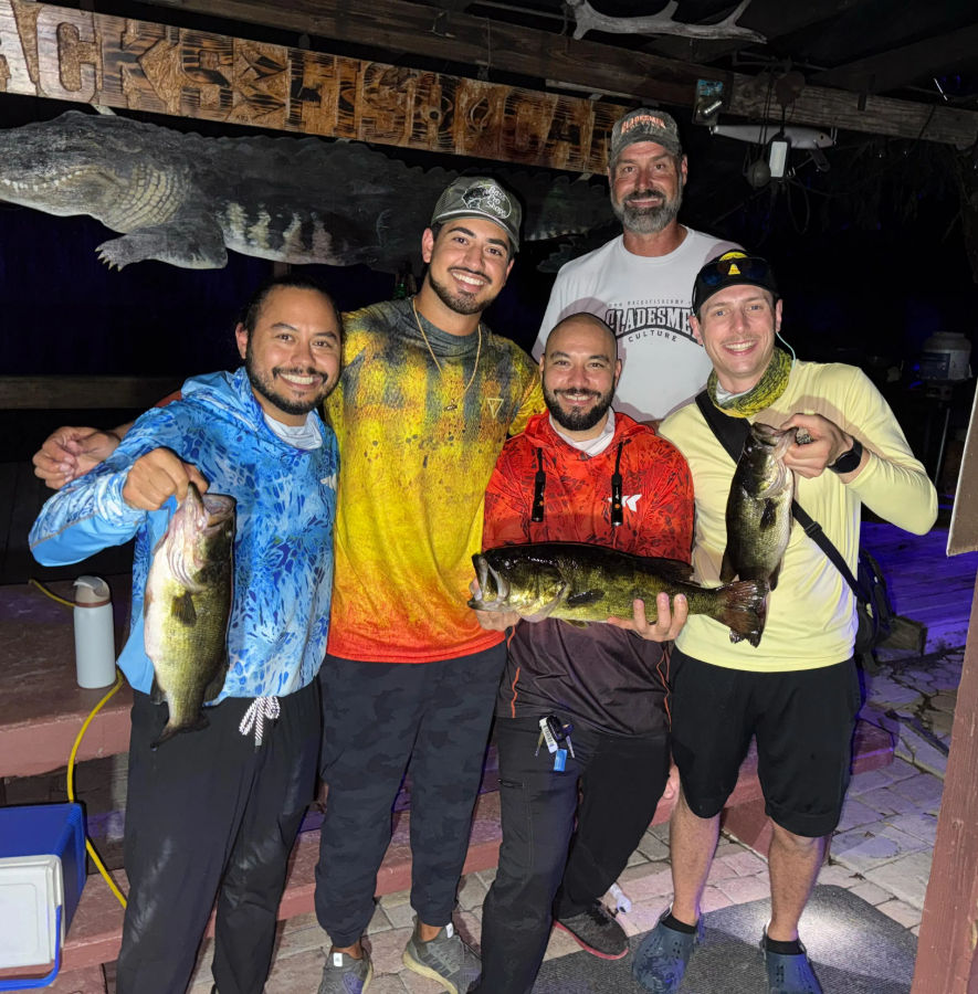 Five smiling anglers on a wooden dock at night holding three large freshwater bass, wearing colorful fishing shirts and celebrating a successful nighttime fishing trip.