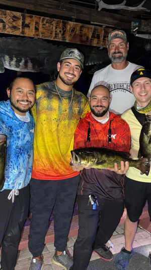 Five smiling anglers on a wooden dock at night holding three freshwater bass, posing under a carved alligator sign after a successful fishing trip