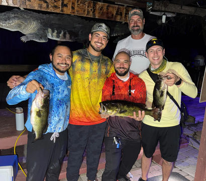 Five smiling anglers on a wooden dock at night holding three freshwater bass, posing under a carved alligator sign after a successful fishing trip