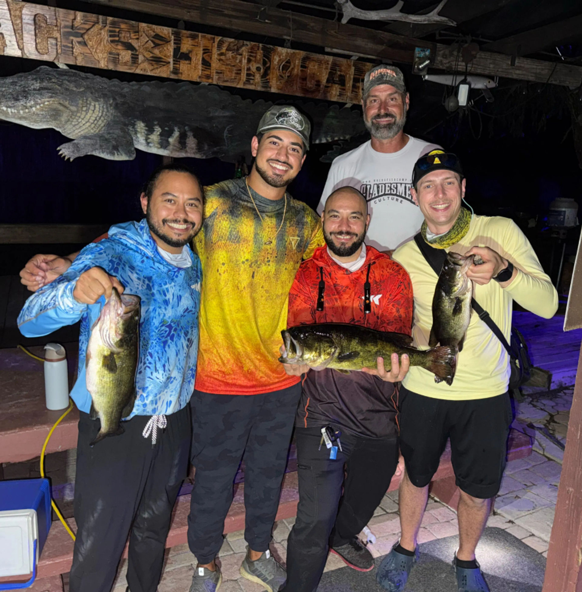 Five smiling anglers on a wooden dock at night holding three freshwater bass, posing under a carved alligator sign after a successful fishing trip