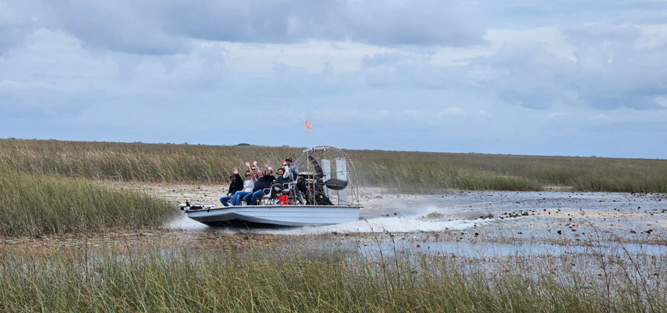 Airboat with passengers waving speeds through shallow marsh water, spraying across grassy Florida Everglades–style wetlands under a cloudy sky