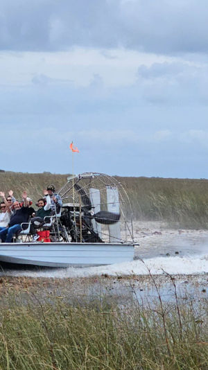 Airboat with passengers waving speeds through shallow marsh water, spraying across grassy Florida Everglades–style wetlands under a cloudy sky