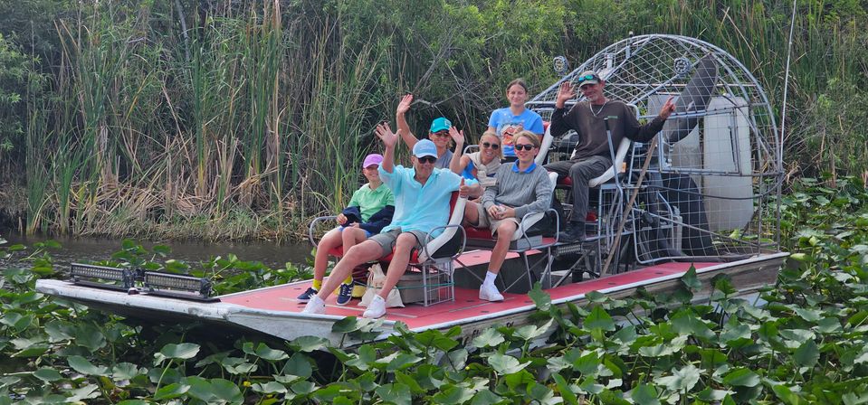 Group of people waving from a red-deck airboat cruising through lily pad–filled wetlands with tall reeds — Florida Everglades airboat tour vibe