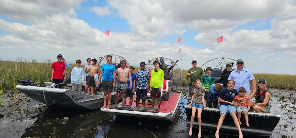 Smiling group of kids and adults standing and sitting on three airboats in the Florida Everglades marsh, muddy feet and wetland grasses under a partly cloudy sky — outdoor airboat adventure.