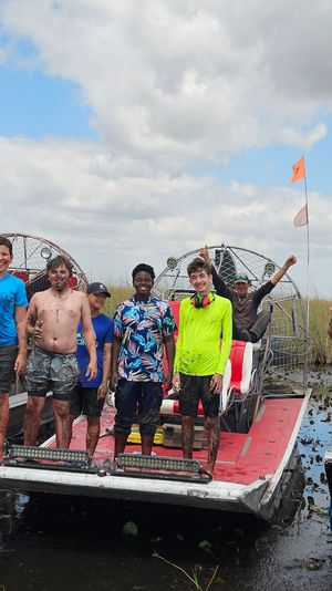 Smiling group of kids and adults standing and sitting on three airboats in the Florida Everglades marsh, muddy feet and wetland grasses under a partly cloudy sky — outdoor airboat adventure.