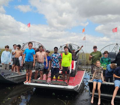 Smiling group of kids and adults standing and sitting on three airboats in the Florida Everglades marsh, muddy feet and wetland grasses under a partly cloudy sky — outdoor airboat adventure.