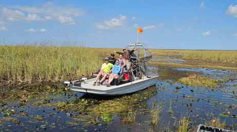 Airboat swamp tour in the Florida Everglades: a small group aboard an airboat navigating shallow marsh with reeds, lily pads and a bright blue sky