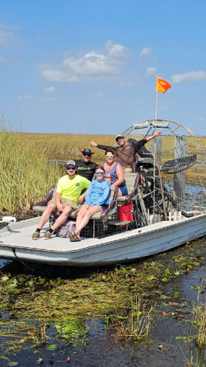 Airboat swamp tour in the Florida Everglades: a small group aboard an airboat navigating shallow marsh with reeds, lily pads and a bright blue sky