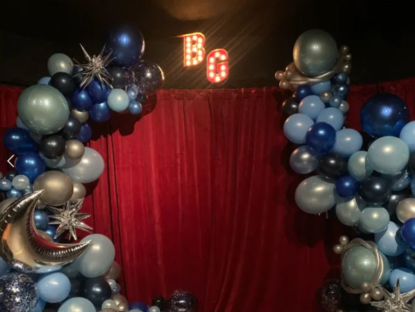 Red velvet photo booth backdrop framed by a blue and silver balloon arch with foil moon and star accents and illuminated 'BG' marquee letters, event décor