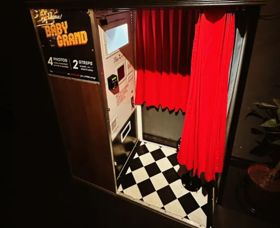 Vintage photo booth interior with bright red curtains, coin-operated control panel and black-and-white checkered floor in a dimly lit indoor venue.
