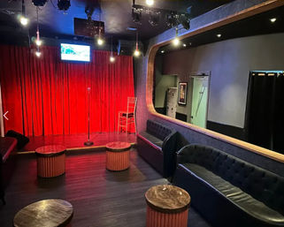 Intimate comedy club interior with small stage framed by red velvet curtains, microphone on a stand and a stool under stage lights, surrounded by tufted black sofas and round wooden cocktail tables.