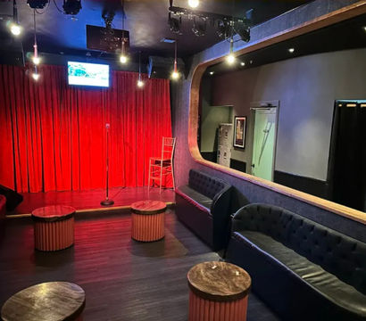 Intimate comedy club interior with small stage framed by red velvet curtains, microphone on a stand and a stool under stage lights, surrounded by tufted black sofas and round wooden cocktail tables.