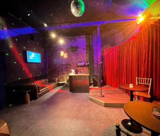 Cozy comedy club interior with a small raised stage and microphone before a red velvet curtain, disco ball and colorful spotlights, exposed brick and compact bar, leather sofa and round cocktail tables.