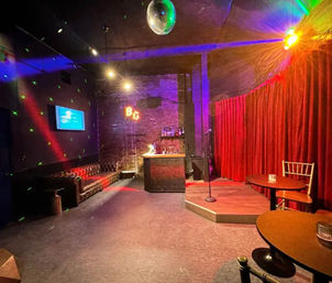 Cozy comedy club interior with a small raised stage and microphone before a red velvet curtain, disco ball and colorful spotlights, exposed brick and compact bar, leather sofa and round cocktail tables.