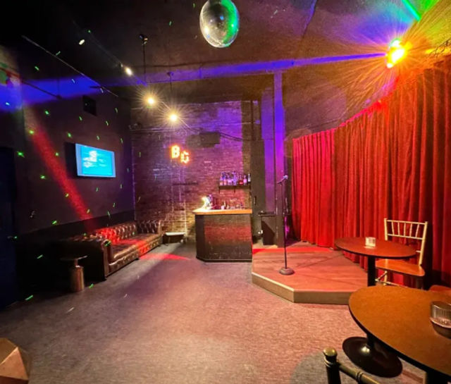 Cozy comedy club interior with a small raised stage and microphone before a red velvet curtain, disco ball and colorful spotlights, exposed brick and compact bar, leather sofa and round cocktail tables.