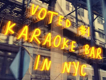 Glowing neon sign reading "Voted #1 Karaoke Bar in NYC" in a storefront window with reflected city buildings and a fire escape, bright urban nightlife vibe.