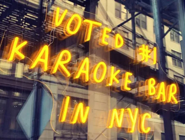 Glowing neon sign reading "Voted #1 Karaoke Bar in NYC" in a storefront window with reflected city buildings and a fire escape, bright urban nightlife vibe.