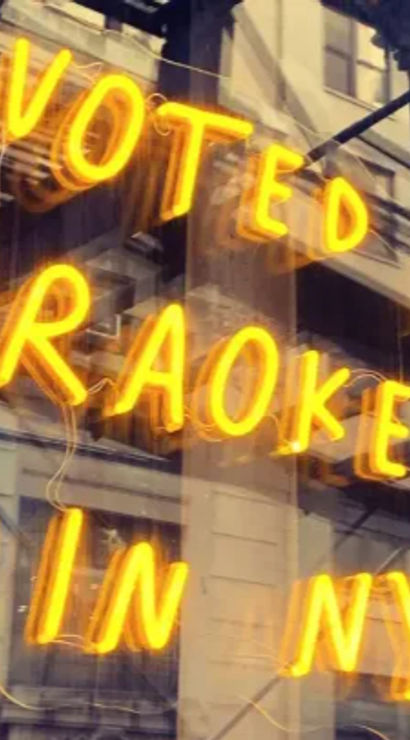 Glowing neon sign reading "Voted #1 Karaoke Bar in NYC" in a storefront window with reflected city buildings and a fire escape, bright urban nightlife vibe.