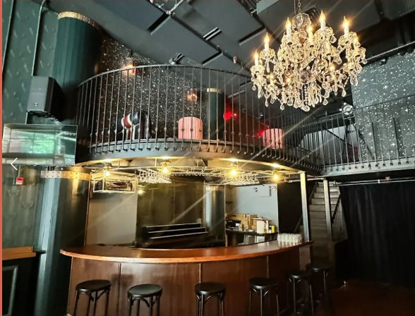 Dimly lit lounge interior with a curved wooden bar and black stools, circular mezzanine balcony above, sparkling crystal chandelier, glitter-speckled black walls and metal staircase — cozy urban nightclub vibe.