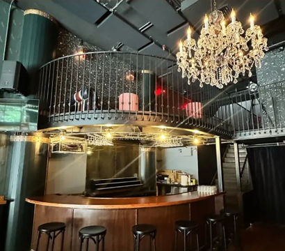 Dimly lit lounge interior with a curved wooden bar and black stools, circular mezzanine balcony above, sparkling crystal chandelier, glitter-speckled black walls and metal staircase — cozy urban nightclub vibe.