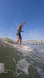 Surfer balancing on a pink longboard riding a small ocean wave near a coastal pier under a clear blue sky