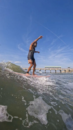 Surfer balancing on a pink longboard riding a small ocean wave near a coastal pier under a clear blue sky