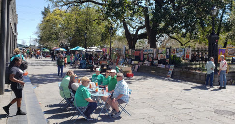 Sunny outdoor art market on a tree-lined paved promenade with artists' paintings hung on an iron fence, street vendors under umbrellas, and a group of people enjoying drinks at small blue bistro tables.