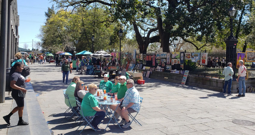 Sunny outdoor art market on a tree-lined paved promenade with artists' paintings hung on an iron fence, street vendors under umbrellas, and a group of people enjoying drinks at small blue bistro tables.