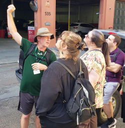 Tour guide in a bucket hat gestures upward while a small group of backpacking visitors look up outside a downtown parking garage on a sunny city street.