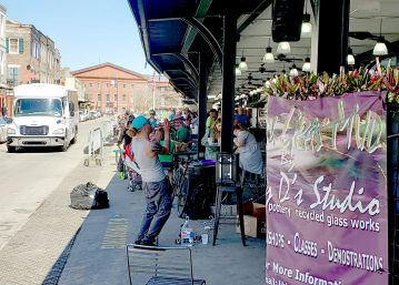 Busker playing trumpet under a covered downtown outdoor market arcade, diners at tables, bicycles and a colorful banner advertising pottery and recycled-glass workshops.