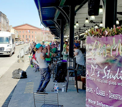 Busker playing trumpet under a covered downtown outdoor market arcade, diners at tables, bicycles and a colorful banner advertising pottery and recycled-glass workshops.