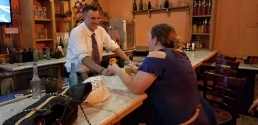 Smiling bartender in a white shirt and tie hands a beer to a woman in a blue cut-out-shoulder dress at a cozy rustic bar with bottles on shelves and a purse on the marble counter.