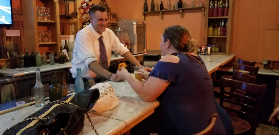 Smiling bartender in a white shirt and tie hands a beer to a woman in a blue cut-out-shoulder dress at a cozy rustic bar with bottles on shelves and a purse on the marble counter.