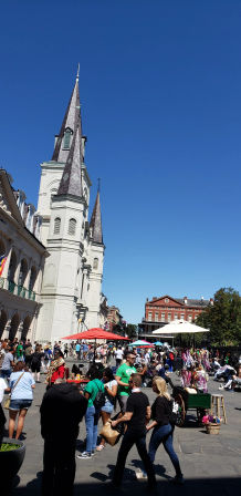 Sunny New Orleans French Quarter scene with St. Louis Cathedral spires towering over a bustling plaza of vendors, colorful umbrellas and crowds of tourists under a clear blue sky.