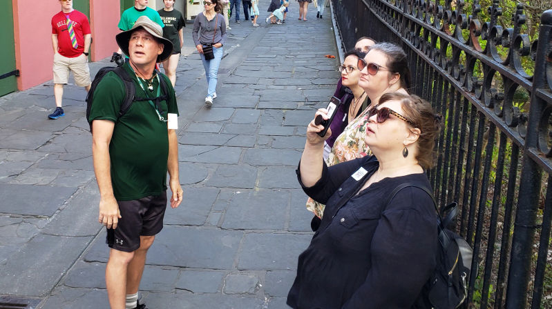Tour group on a historic stone-paved street by an ornate iron fence, man in green shirt and bucket hat looking up while a woman in sunglasses snaps a photo with her phone — urban walking tour scene.