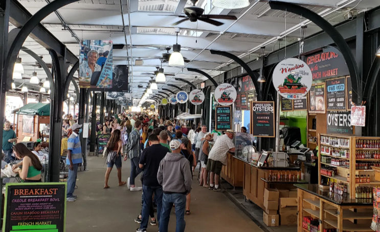 Bustling covered market pavilion with arched metal beams, long aisle of seafood and food stalls (oysters, sauces), hanging lights and ceiling fans, crowds of shoppers queued and browsing.