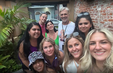 Smiling group selfie of nine friends on a brick-walled outdoor patio with tropical plants, sunglasses, and colorful summer outfits.