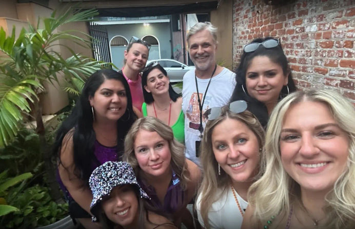Smiling group selfie of nine friends on a brick-walled outdoor patio with tropical plants, sunglasses, and colorful summer outfits.