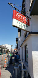 Vintage red soda sign hanging over a narrow downtown sidewalk cafe with metal bar stools, pedestrians and a palm tree under a bright blue sky
