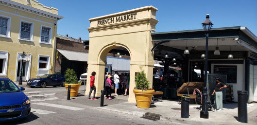 Sunny street scene with a beige arched market entrance to an outdoor market, shoppers gathered under the arch, large yellow planters, open-air stalls under a canopy, historic-style buildings and parked cars nearby.