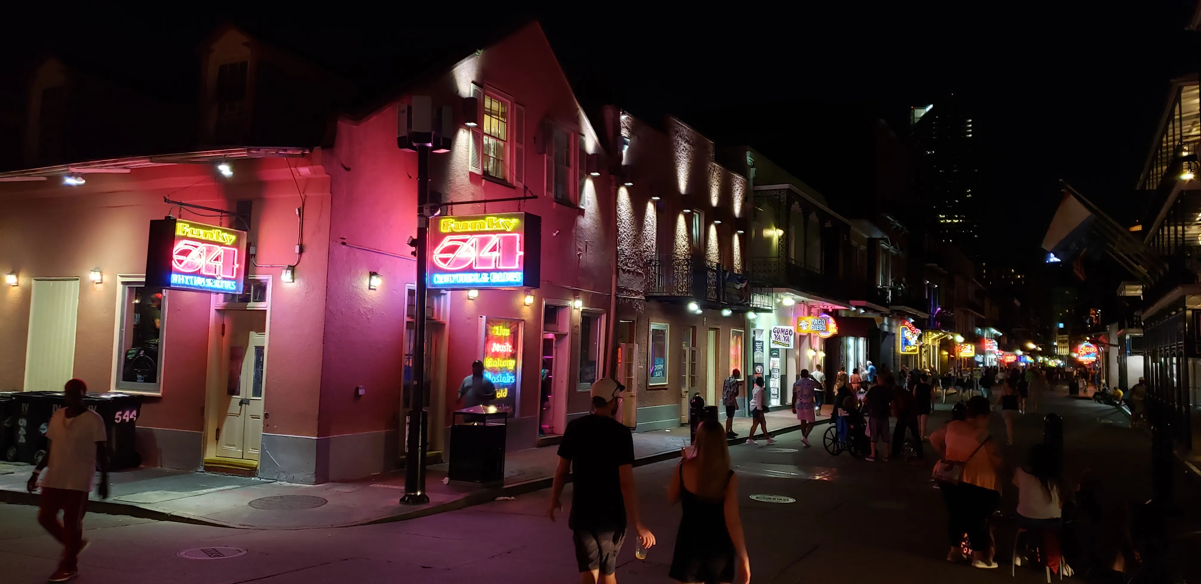 Nighttime New Orleans French Quarter street scene with neon bar signs and pink‑lit historic buildings, crowds of pedestrians and a lively nightlife vibe.