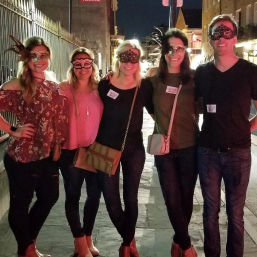 Five friends wearing decorative masquerade masks posing together on a lively historic downtown street at night, casual outfits and crossbody bags in a nightlife scene.