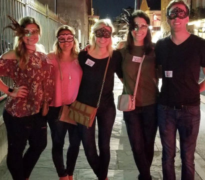 Five friends wearing decorative masquerade masks posing together on a lively historic downtown street at night, casual outfits and crossbody bags in a nightlife scene.