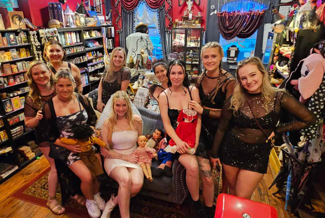 Smiling group of women posing inside a quirky vintage gift shop—shelves of books, curios and dolls surround a woman in a white dress and veil seated on a couch, friends in black party outfits holding vintage dolls under warm red-and-black decor.