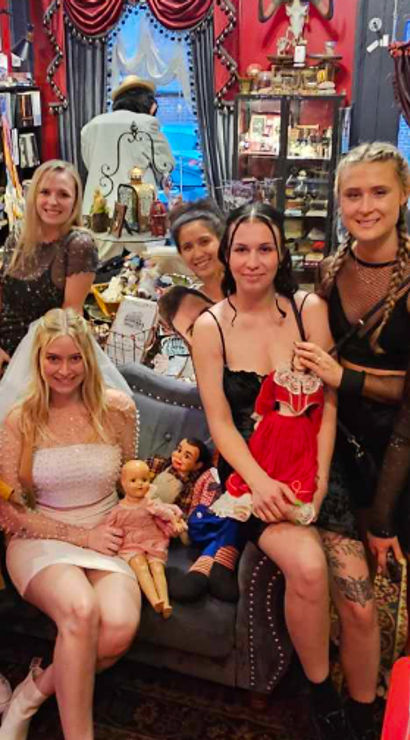 Smiling group of women posing inside a quirky vintage gift shop—shelves of books, curios and dolls surround a woman in a white dress and veil seated on a couch, friends in black party outfits holding vintage dolls under warm red-and-black decor.